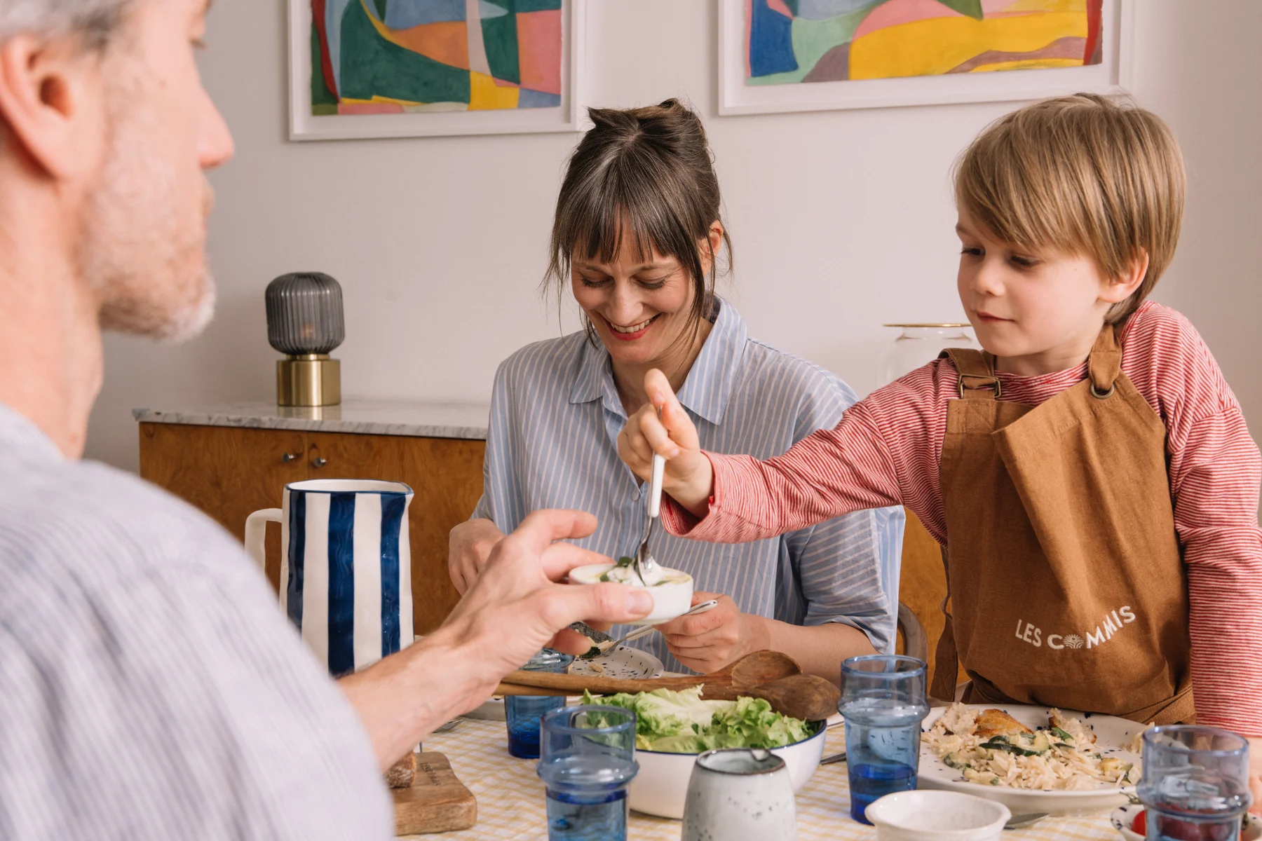 Une famille partage un repas à table, l'enfant porte un tablier Les Commis et sert une assiette