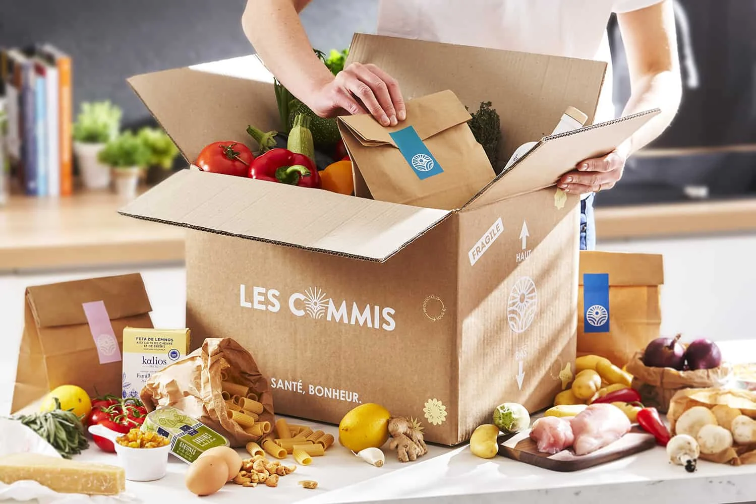 Une personne ouvre un carton Les Commis rempli de légumes frais et de sachets d'ingrédients dans sa cuisine