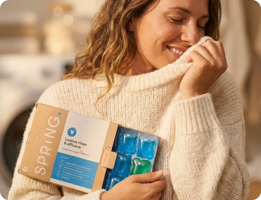 Une femme respire l'odeur de son pull propre en tenant une boîte de lessive Spring